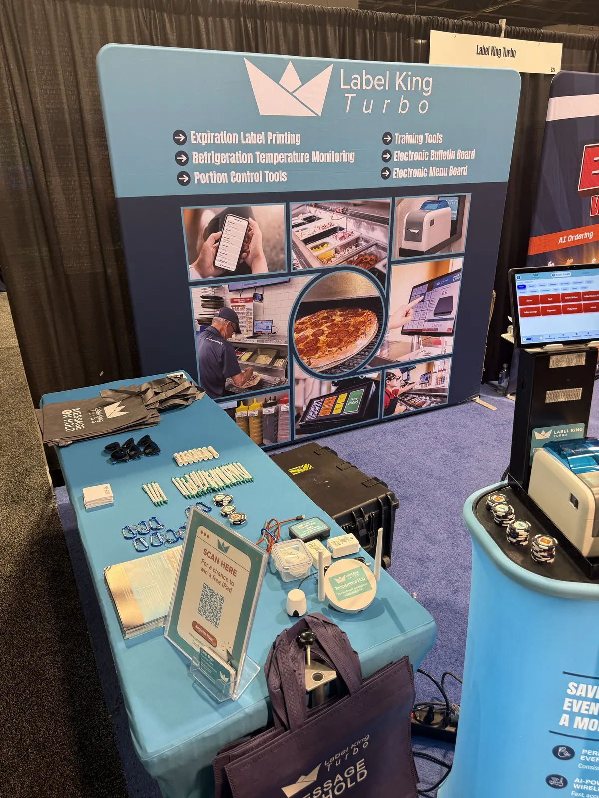 Label King Turbo booth table stocked with giveaways, wireless temperature sensors, and a QR-code sign in front of the show-floor backdrop